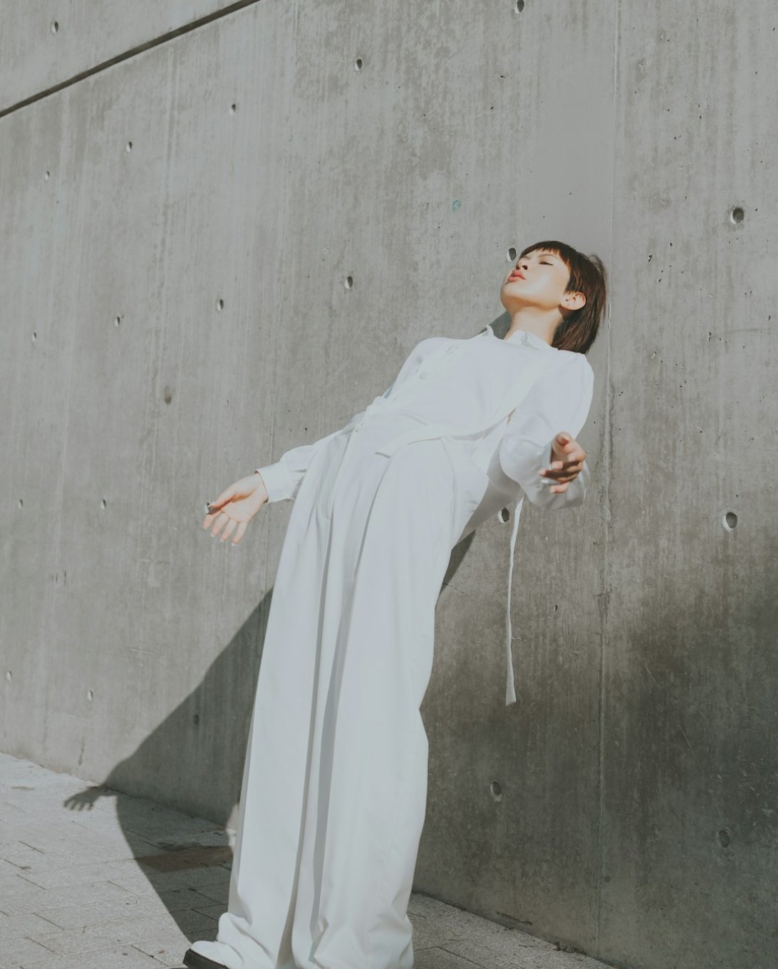 A model in a loose white outfit leaning back against a concrete wall